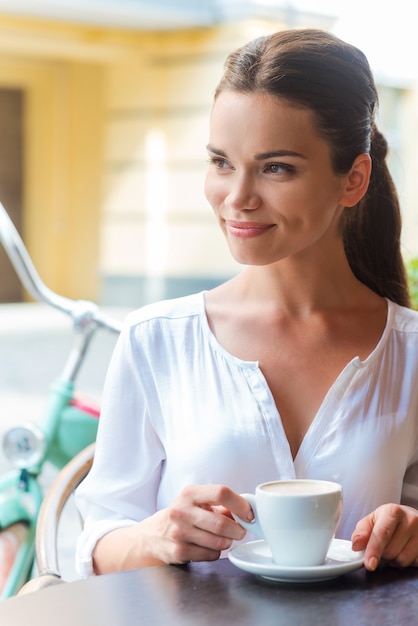 Relaxar com uma xícara de café fresco. Mulher jovem e bonita tomando café e olhando para longe enquanto está sentada na calçada do café