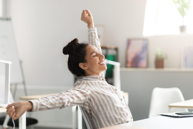 Foto relaxe no trabalho feliz jovem gerente sentada no escritório esticando as costas e os braços sentindo-se satisfeita e satisfeita