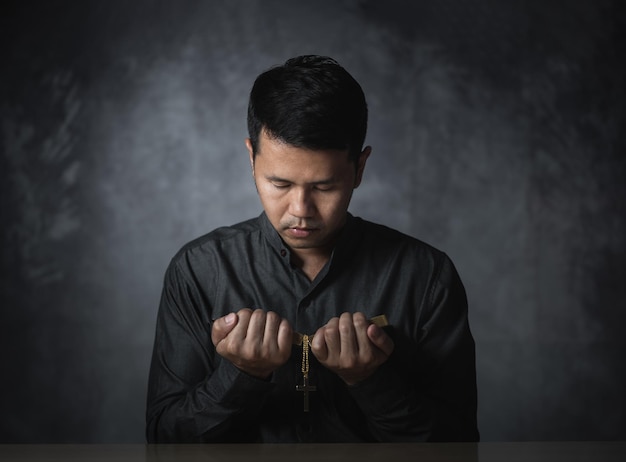 Religiöser asiatischer Mann, der mit einer Halskette aus Kreuzen und Bibelversen betet, die für heilige Segnungen beten, die auf dem grauen Hintergrund des Tisches sitzen Spiritualität und Religion Christliches Gebetsreligionskonzept