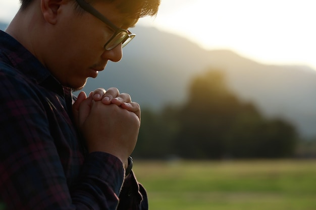 Religiöser Mann, der zu Gott betet, während eines schönen Sonnenuntergangs sein Kinn auf seinen Händen auf einem Feld ruht