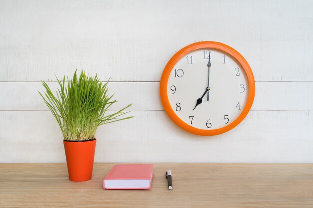 Relógio de parede redondo, bloco de notas rosa sobre a mesa e planta de casa verde. Ambiente de trabalho. Início da jornada de trabalho