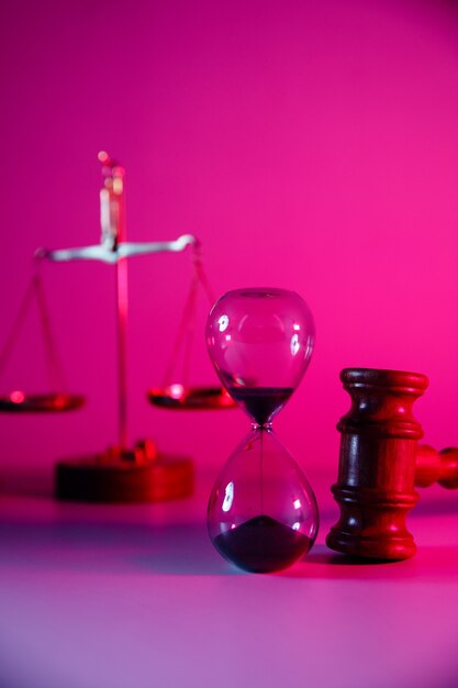 Reloj de arena, balanza y mazo de juez de madera en neón rosa. Imagen vertical.