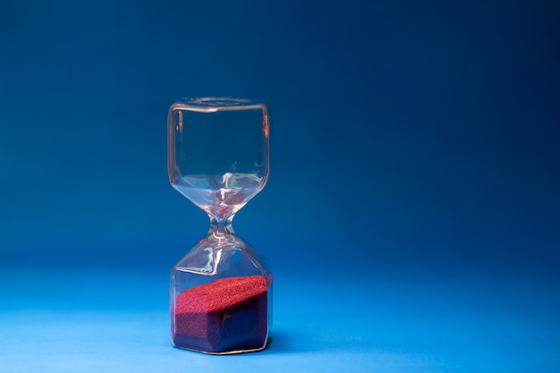 Reloj de arena transparente con granos rojos de arena ubicados sobre un fondo azul con espacio libre para texto