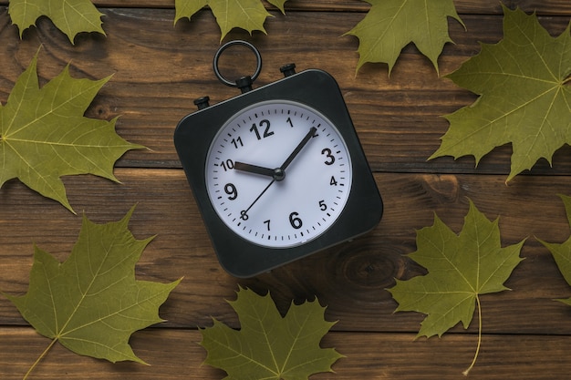 Reloj blanco y negro con manecillas y hojas de arce sobre un fondo de madera.