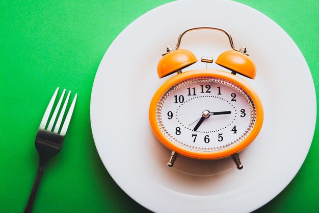 Reloj despertador de estilo vintage naranja en un plato blanco con un tenedor Concepto de tiempo de comida simple plano
