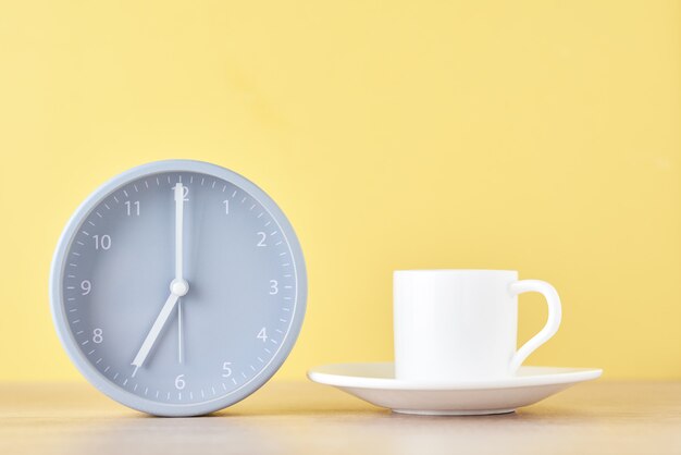 Reloj despertador gris clásico y taza de café con leche sobre un fondo amarillo