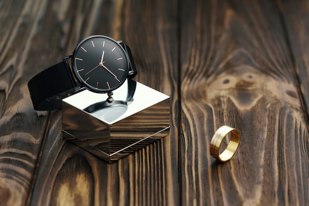 Reloj moderno negro en cubo cromado con anillo de oro sobre fondo de madera