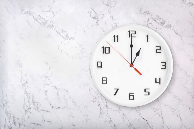 Reloj de pared redondo blanco sobre fondo de mármol blanco natural. La una en punto