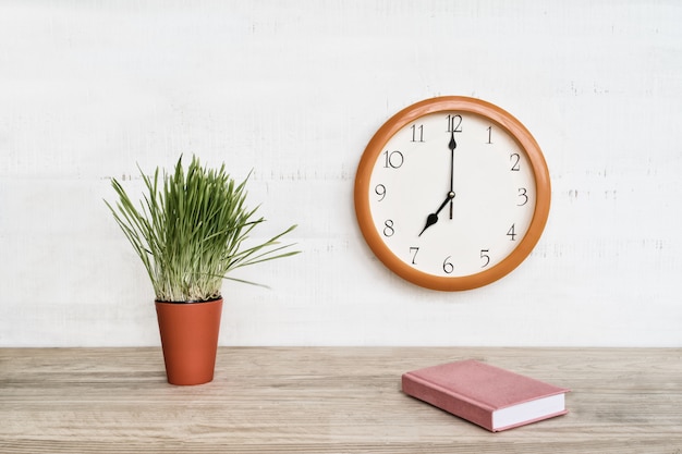 Reloj de pared redondo, libreta rosa sobre la mesa y planta de interior verde