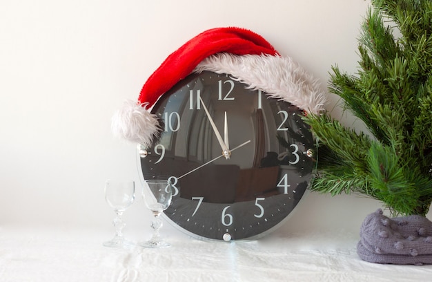 Un reloj con un sombrero de santa claus junto a un árbol de navidad artificial y dos vasos.