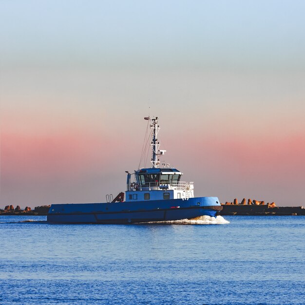 Remolcador pequeño azul navegando más allá de la presa del rompeolas en la mañana