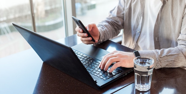 Remote-Arbeitsplatz. Freiberufler-Arbeitsplatz in der Nähe des Fensters. Mann, der Laptop verwendet, Smartphone für geschäftliche Arbeit hält oder in Smartwatch Wasser trinkt. Langes Webbanner.