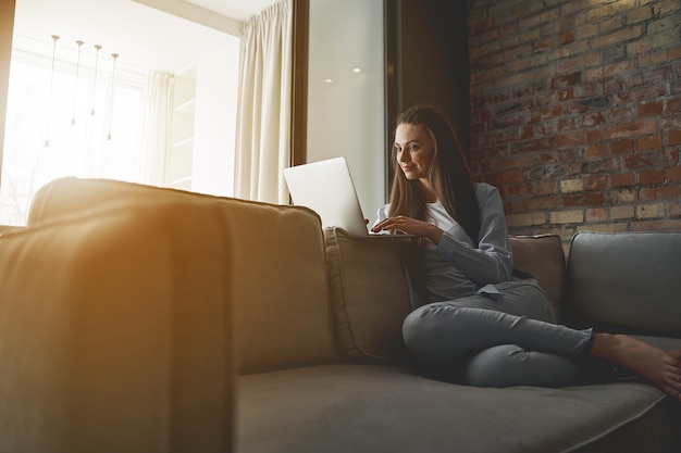 Remote-Arbeitsplatz. Schöne lächelnde fokussierte junge Frau in Freizeitkleidung, die drinnen auf ihrem Laptop tippt