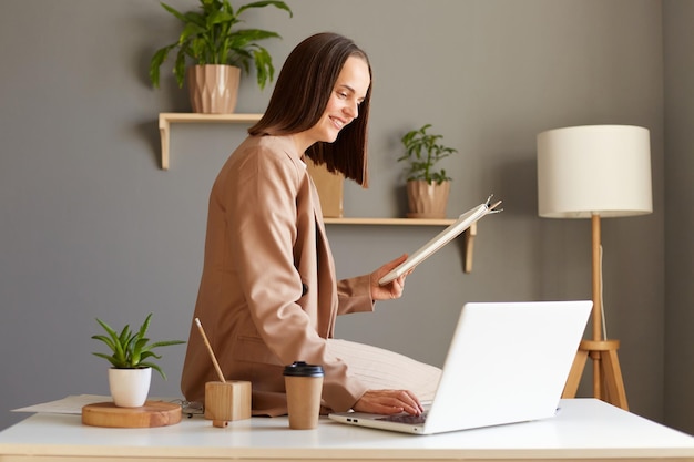 Remote-Job-Technologie und Menschen Porträt einer glücklich lächelnden kaukasischen jungen Frau mit Laptop-Computer und Papieren in der Zwischenablage, die zu Hause im Büro arbeitet und eine Jacke trägt, die auf dem Tisch sitzt