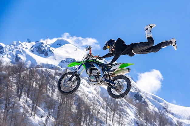 Rennfahrer auf einem Motorrad im Flug, springt und startet auf einem Sprungbrett gegen die schneebedeckten Berge