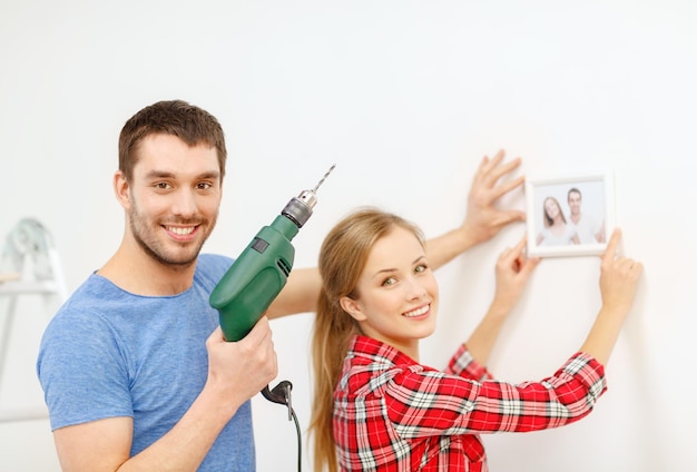 reparação, design de interiores, construção, renovação e conceito de casa - casal sorridente fazendo furo na parede e colocando foto em casa