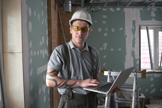 Reparação do quarto. construtor de capacete e óculos fica com um laptop nas mãos no fundo da construção