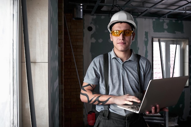 Reparação do quarto. Construtor de capacete e óculos fica com um laptop nas mãos no fundo da construção