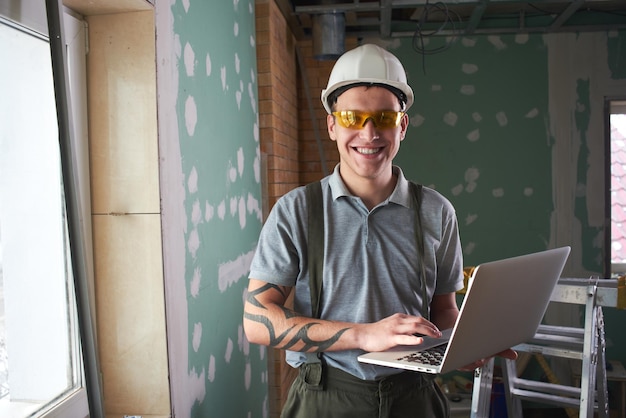 Reparação do quarto. Sorridente jovem construtor de capacete e óculos fica com um laptop na mão no contexto de um canteiro de obras