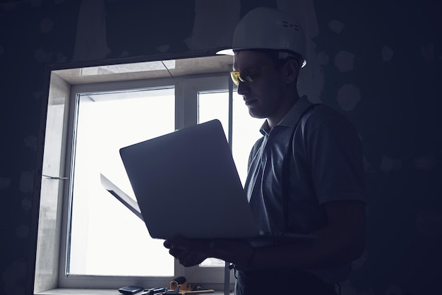 Reparación de habitaciones. Constructor con casco y anteojos con una laptop y dibujos en sus manos en el fondo de la construcción