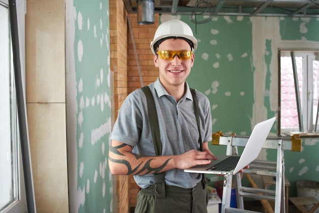 Foto reparación de habitaciones. constructor con casco y anteojos parados con una laptop en sus manos en el fondo de la construcción