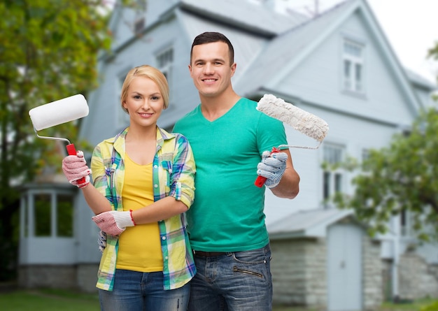 reparación, personas, bienes raíces, hogar y concepto familiar - pareja sonriente con rodillos de pintura sobre el fondo de la casa