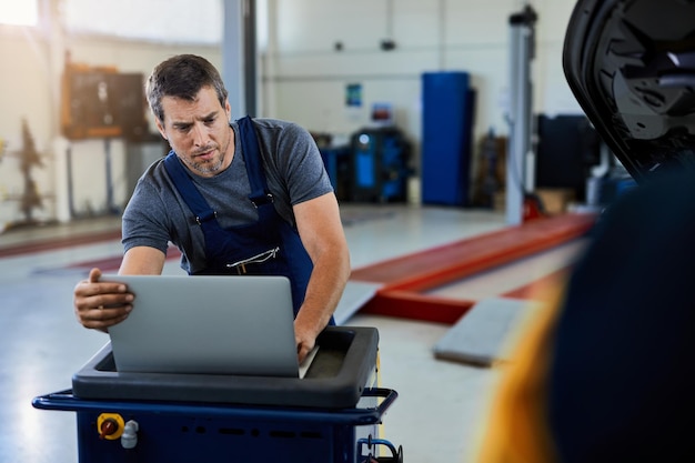 Reparador de automóviles que trabaja en una computadora portátil mientras ejecuta un diagnóstico de automóvil en un taller