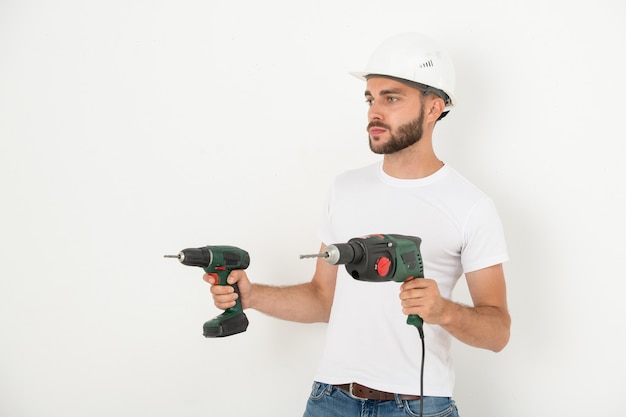 Reparador barbudo joven serio en casco sosteniendo herramientas eléctricas de mano mientras se prepara para remodelar en piso