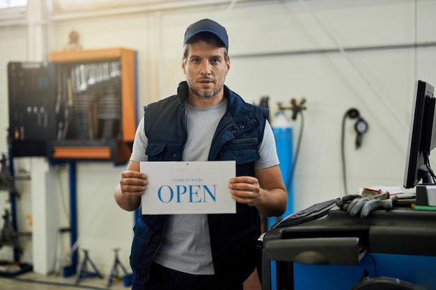 Reparador de automóveis segurando placa aberta enquanto está em uma oficina e olhando para a câmera