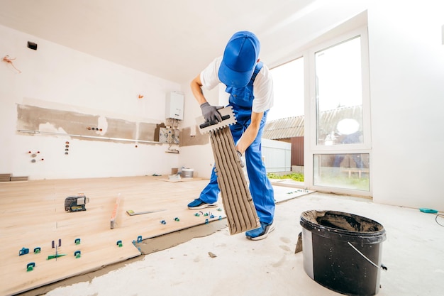 Reparador con guantes grises y uniforme especial que pone cemento sobre baldosas en una casa nueva