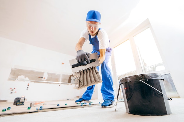 Reparador con guantes grises y uniforme especial que pone cemento sobre baldosas en una casa nueva