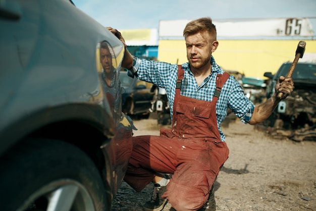 Reparador masculino trabaja con un martillo en el depósito de chatarra del coche. Chatarra de automóviles, chatarra de vehículos, basura de automóviles, transporte abandonado, dañado y aplastado