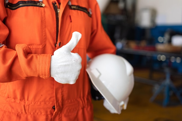 Foto un reparador en mono naranja está levantando el pulgar y sosteniendo un casco de seguridad blanco