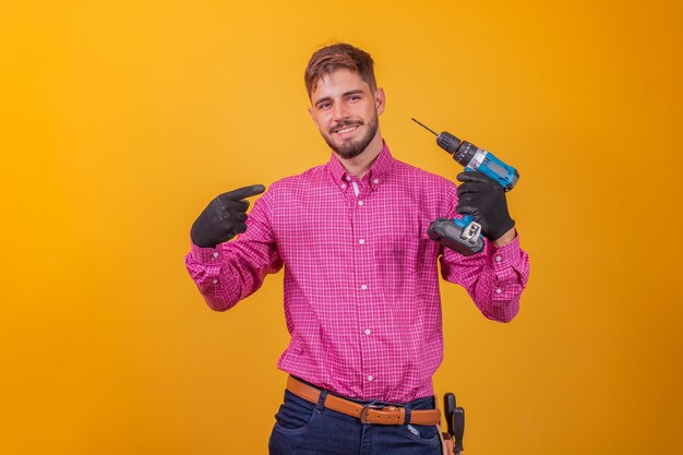 Reparador positivo e confiante em uma camisa xadrez e um kit de ferramentas no quadril e uma chave de fenda elétrica sorrindo e olhando para a câmera
