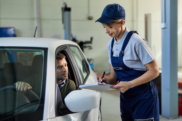 Reparadora de automóviles escribiendo datos mientras habla con su cliente que está sentado en un automóvil en un taller de reparación de automóviles