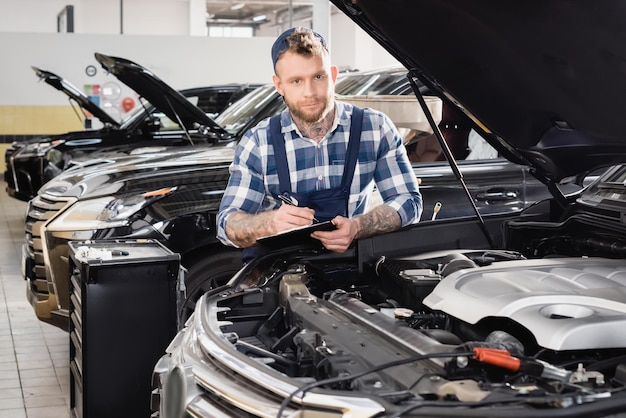 Foto reparadores sosteniendo el clipboard y mirando a la cámara cerca del compartimento del motor del coche