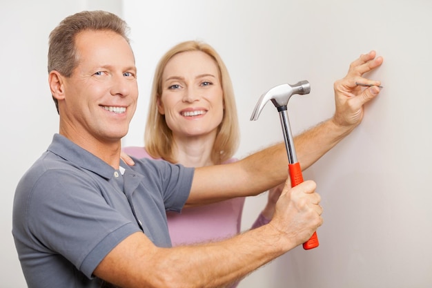 Reparando casa juntos. Hombre de pelo gris alegre martillando un clavo y sonriendo mientras su esposa está de pie cerca de él