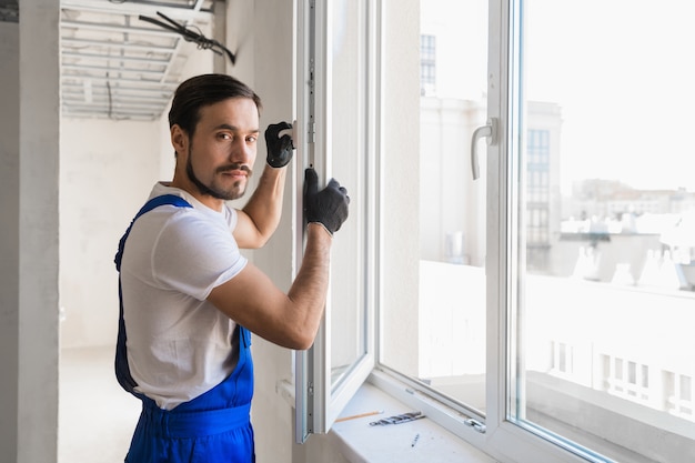 Reparateur in Overalls baut am Fenster und schaut in die Kamera