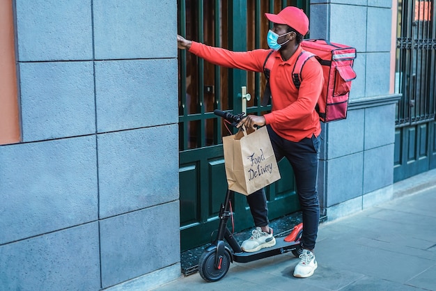 Repartidor africano con scooter eléctrico tocando el timbre - Centrarse en la bolsa de papel