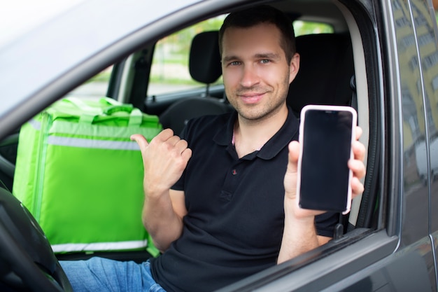 El repartidor de alimentos tiene una mochila nevera verde. Se sienta en un carro. Muestra la pantalla de un teléfono inteligente con una aplicación de entrega de alimentos.