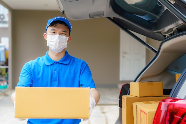 El repartidor asiático atiende a un mensajero que trabaja con cajas de cartón en una camioneta durante la pandemia del coronavirus (COVID-19), un mensajero con máscara médica y guantes de látex por seguridad.