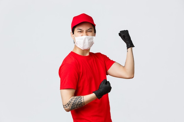 Repartidor asiático entusiasta con gorra y camiseta de uniforme rojo, guantes protectores y máscara médica, aumentando la confianza, bomba de puño, celebrando el éxito, listo para transferir paquetes a los clientes.