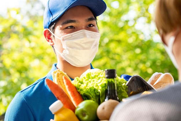 Repartidor asiático higiénico con mascarilla facial mientras entrega comestibles al cliente en casa, entrega de alimentos en el momento del concepto de pandemia