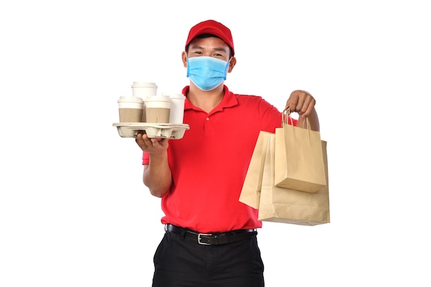 Repartidor asiático joven feliz en uniforme rojo, mascarilla médica llevar bolsas de comida y bebida en manos aisladas sobre fondo blanco