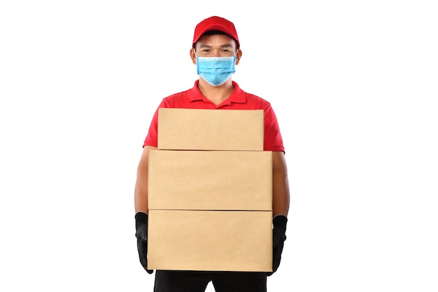 Repartidor asiático con mascarilla y guantes en uniforme rojo entregando caja de paquetería aislada en blanco durante el brote de COVID-19