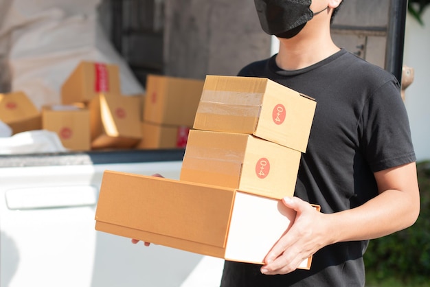 Repartidor asiático con mascarilla en uniforme negro sosteniendo cajas de cartón en frente de la casa