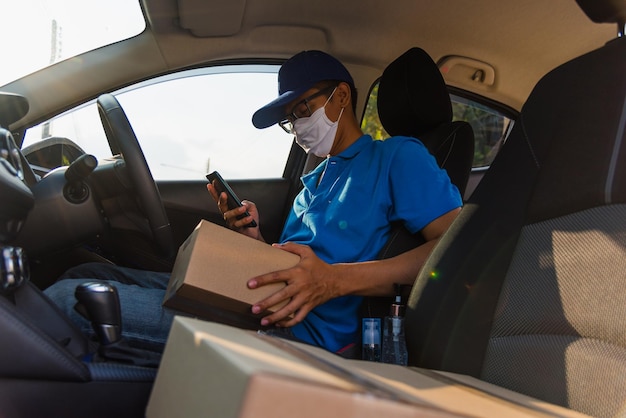 Repartidor asiático repartidor en línea que realiza entregas en cajas en el automóvil y usa el teléfono móvil para contactar al cliente con el servicio de mascarilla protectora bajo toque de queda cuarentena pandemia coronavirus COVID-19