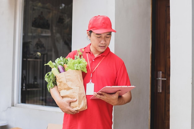 Repartidor asiático revisando el documento en el portapapeles mientras maneja una bolsa de verduras.