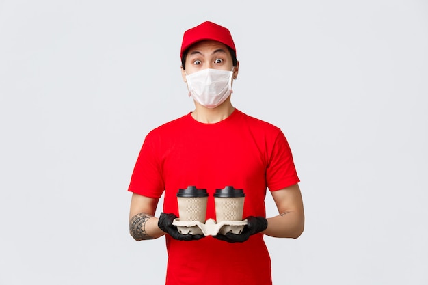 Repartidor asiático sorprendido con guantes y mascarilla médica, mirando al cliente, trayendo una persona encargada del pedido de comida de café, ordenó la aplicación móvil en línea durante el brote de covid-19, el mensajero se sorprendió