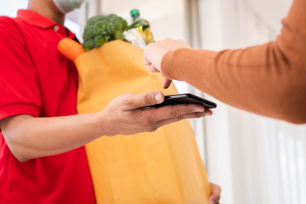 Repartidor asiático sosteniendo una bolsa de comida fresca para dársela a los clientes y sosteniendo un teléfono inteligente para recibir pagos en casa